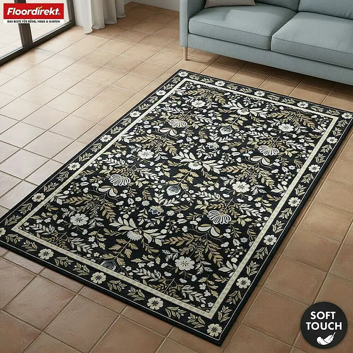 Floordirekt Kurzflorteppich Aristo 160 x 230 cm - Schwarz, 230 x 160 cm - Ein weicher schwarzer Teppich mit komplexem weiß-goldenen Blumenmuster unter einem hellblauen Sofa auf Fliesen.