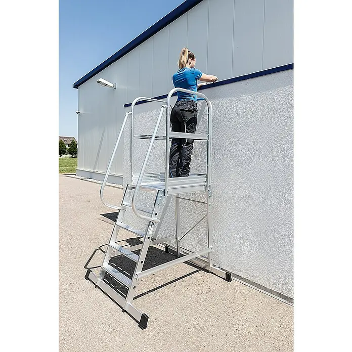Proregal Stehleiter Plattformleiter mit Rollen - Anzahl Tritte: 4 Stk., Arbeitshöhe: 2.950 mm, Belastbarkeit: 150 kg, Aluminium - Aluminium-Arbeitsplattform mit Treppe und Handläufen, eine Frau arbeitet damit an einer Gebäudewand.