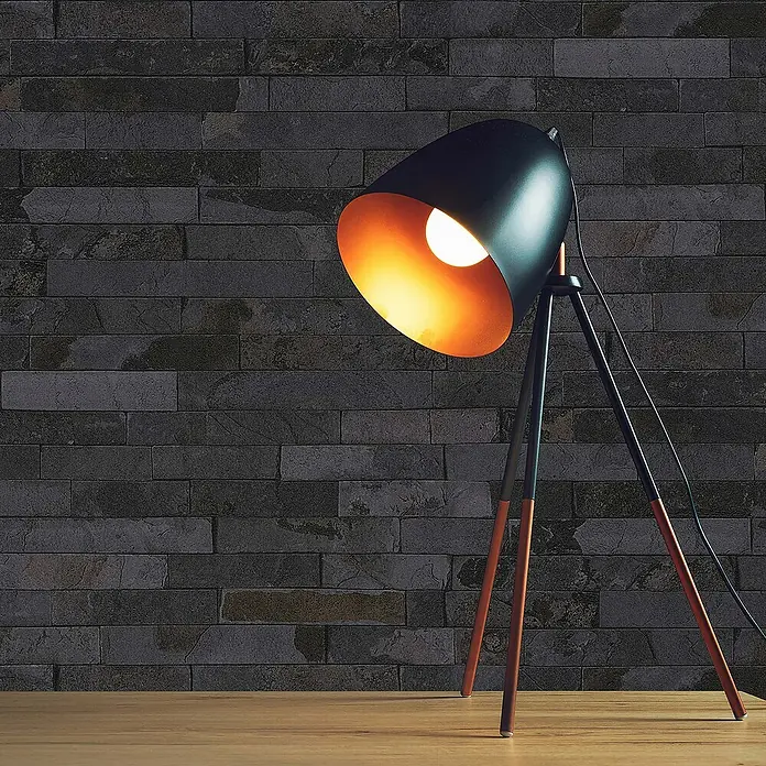 Tadessi Vliestapete M1-No.112 - Schwarz, Steinoptik, 10,05 x 0,53 m - Schwarze und kupferne Stativ-Schreibtischlampe mit warmem Licht auf Holztisch vor dunkler Ziegelwand.