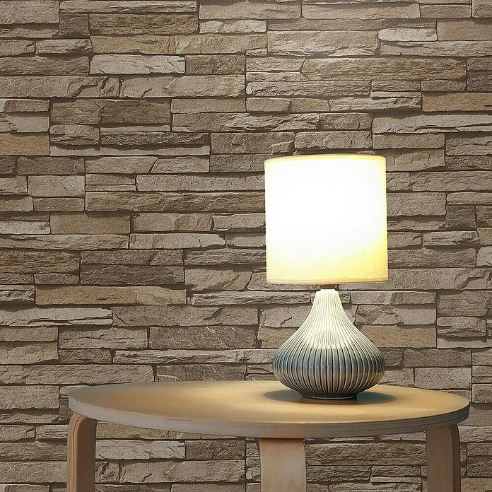 Tadessi Vliestapete M1-No.3817 - Braun, Steinoptik, 10,05 x 0,53 m - Eine beleuchtete Tischlampe mit hellem Schirm und geripptem dunklem Sockel auf einem runden Holztisch vor einer Steinwand.