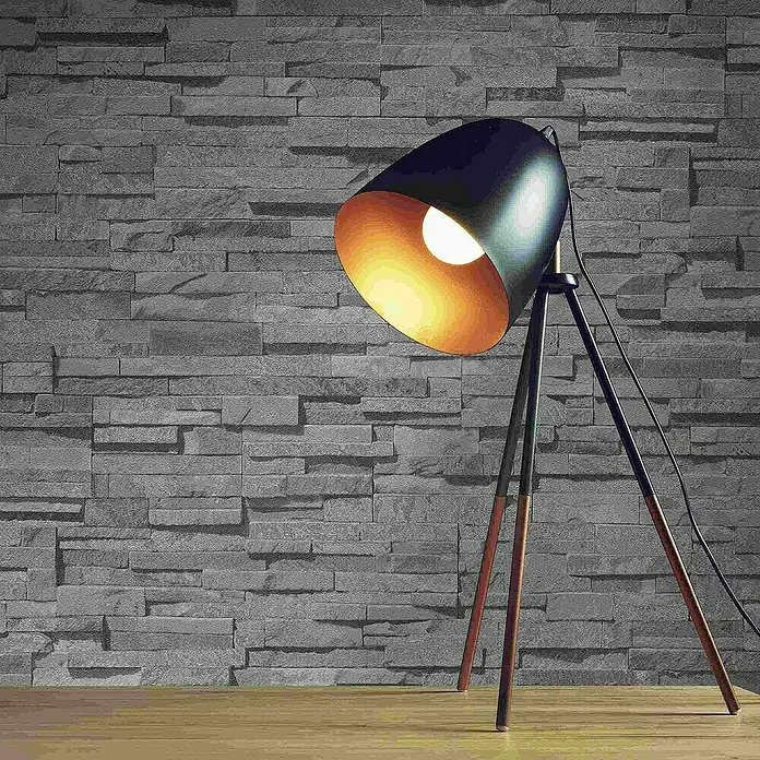 Tadessi Vliestapete M1-No.4322 - Grau, Steinoptik, 10,05 x 0,53 m - Schwarze Stehlampe mit Holzbeinen und goldener Innenseite des Schirms, leuchtet auf eine Ziegelwand.