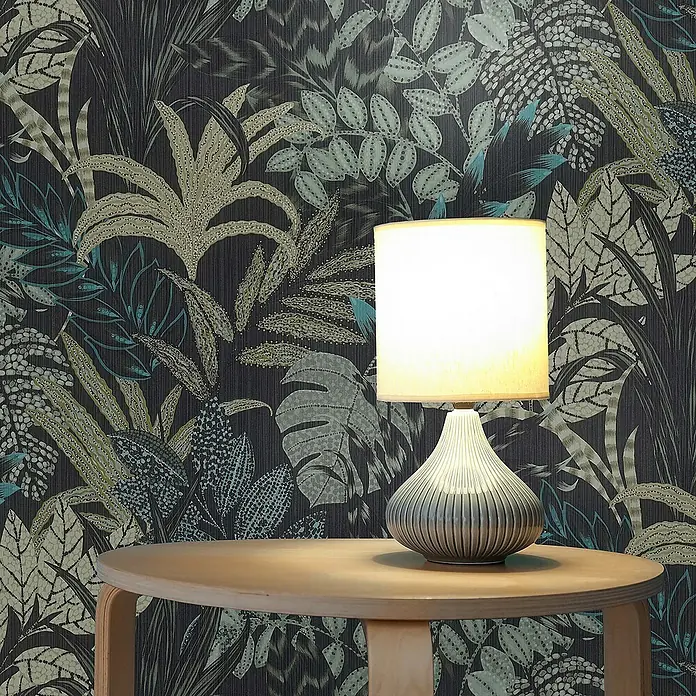 Tadessi Vliestapete M1-No.609 - Schwarz, Floral, 10,05 x 0,53 m - Graue geriffelte Tischlampe mit hellem Schirm steht auf Holztisch und beleuchtet dunkle Tropen-Tapete.