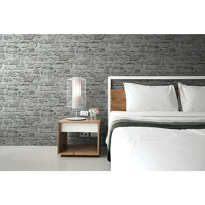 Tadessi Vliestapete M1-No.185 - Grau, Steinoptik, 10,05 x 0,53 m - Schlafzimmeransicht mit weißem Bett, Holzkopfteil, Nachttisch und weißer Lampe vor einer grauen Steinwand.