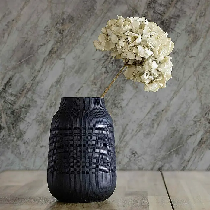Tadessi Vliestapete M1-No.4011 - Schwarz, Steinoptik, 10,05 x 0,53 m - Dunkelblaue Vase mit gerippter und gerasterter Textur, die eine getrocknete Hortensie auf einem Holztisch hält.