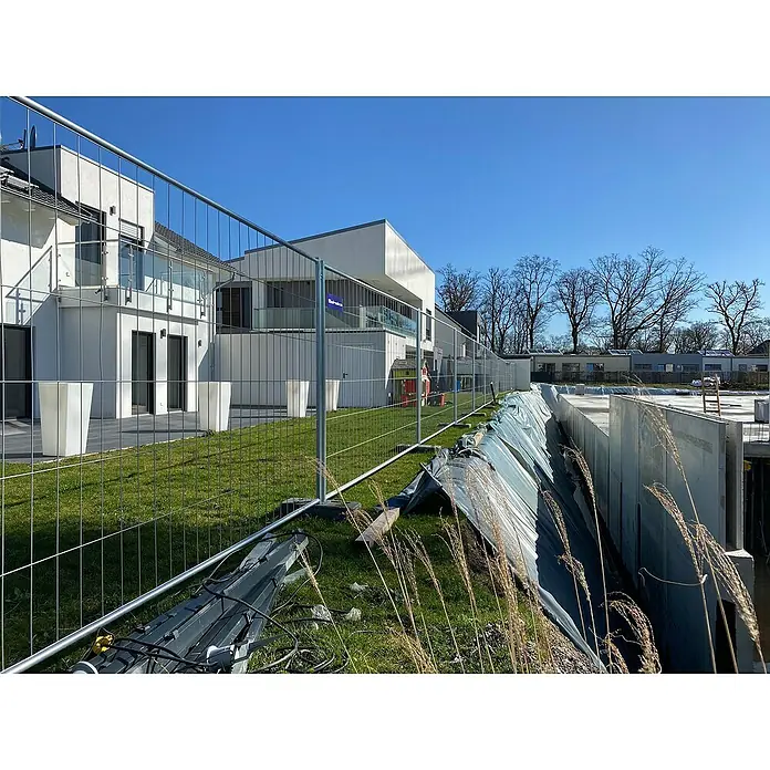 Schake Bauzaun Set Standard - Länge: 35 m, Höhe: 2 m, Stahl - Baustellenszene mit modernen Gebäuden hinter einem Bauzaun. Im Vordergrund Baugrube mit Schutzfolie und Gräsern.