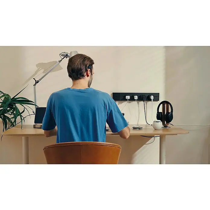 Schwaiger Steckdosenleiste Modulare Stromschiene - 50 cm, Schwarz, IP20, Ohne Schalter - Person, die an einem Schreibtisch mit einer Steckdosenleiste an der Wand arbeitet. Auf dem Tisch stehen Kopfhörer und ein Laptop.