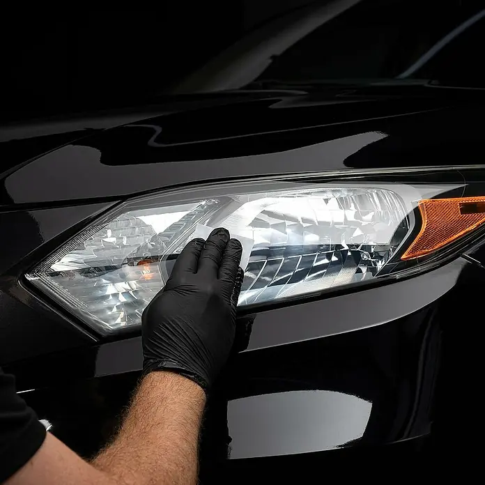 Cerakote Scheinwerfer-Aufbereitungs-Set - 15 -tlg. - Scheinwerferreinigung eines schwarzen Autos mit einer Hand mit schwarzem Handschuh.