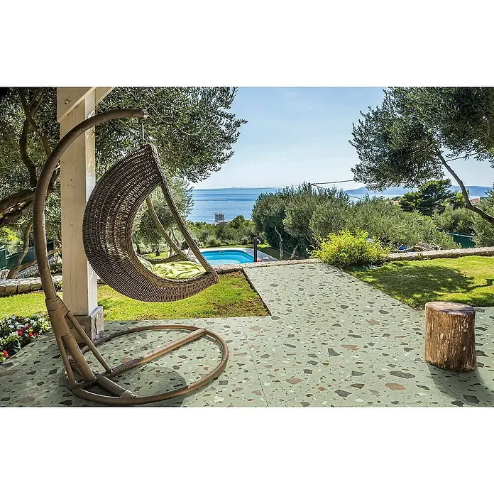 Feinsteinzeugfliese Pebble - 59,55 x 59,55 cm, Green, Matt - Ein schwebender Rattansessel mit grünem Kissen hängt auf einer grünen Terrasse mit Blick auf das Meer.