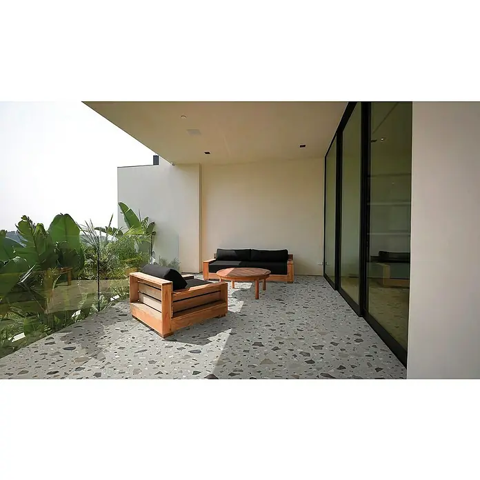 Feinsteinzeugfliese Pebble - 59,55 x 59,55 cm, Grey, Matt - Terrassenmöbel-Set mit Sofa, Sessel und Tisch auf stilvollem Terrazzoboden, umgeben von üppigem Grün und modernen Glasschiebetüren.