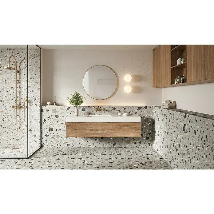 Feinsteinzeugfliese Pebble - 59,55 x 59,55 cm, Grey, Matt - Ein Badezimmer mit Terrazzoboden, Waschtisch und Dusche. Die goldenen Armaturen und der runde Spiegel verleihen dem Raum Eleganz und Stil.