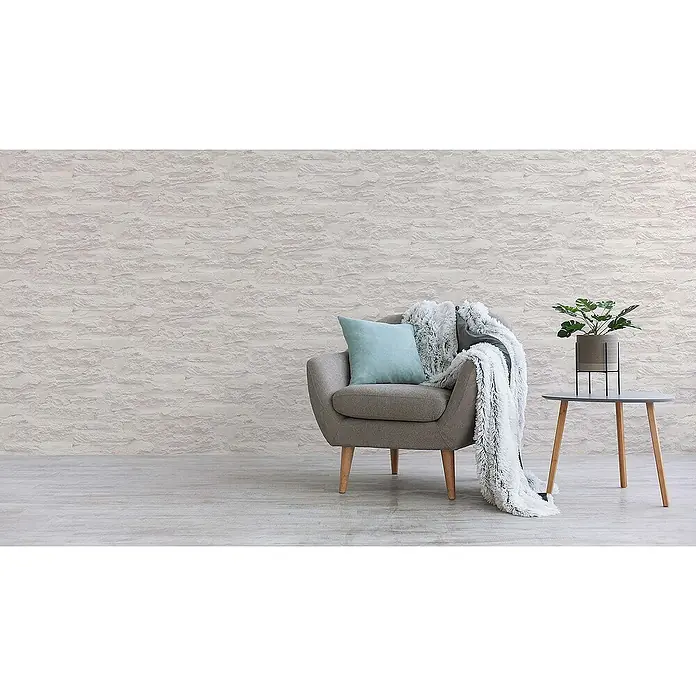 Tadessi Vliestapete M1-No.913 - Creme, Steinoptik, 10,05 x 0,53 m - Strukturierte hellgraue Tapete an einer gemütlichen Wohnzimmerwand hinter einem Sessel mit Kissen und Decke.