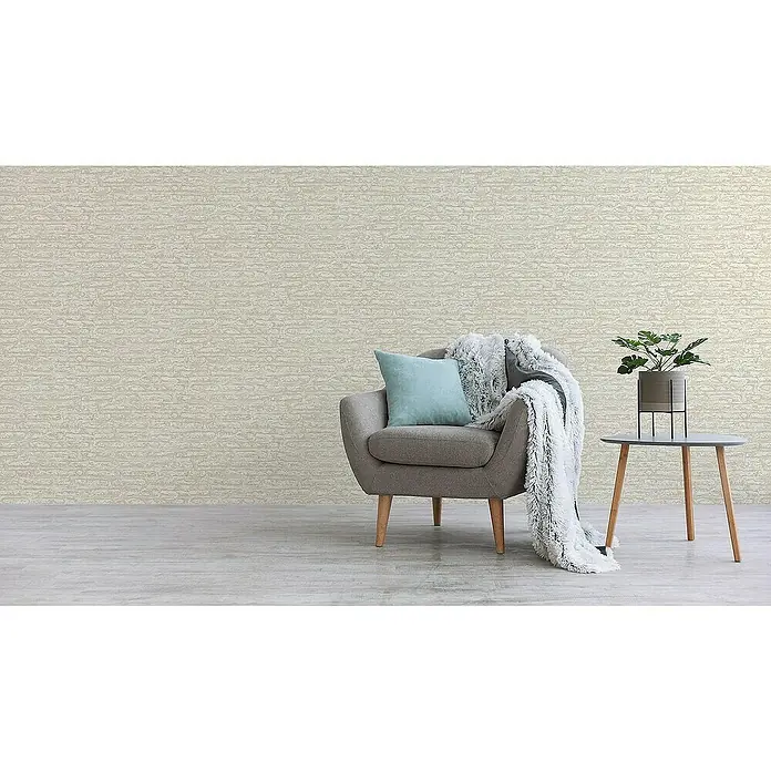 Tadessi Vliestapete M1-No.4192 - Weiß, Steinoptik, 10,05 x 0,53 m - Beigefarbene strukturierte Tapete an einer Wand hinter einem grauen Sessel mit blauem Kissen und Decke, und einem Beistelltisch mit Pflanze.