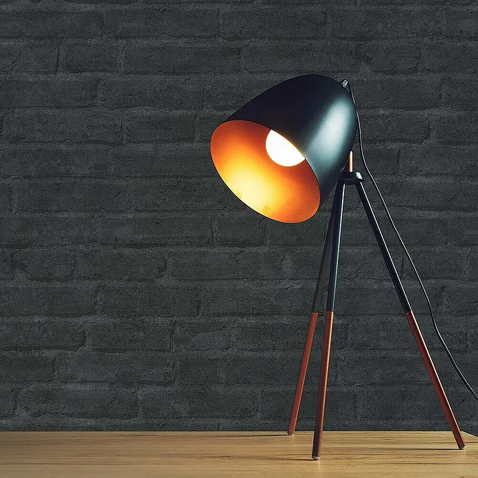 Tadessi Vliestapete M1-No.1415 - Schwarz, Steinoptik, 10,05 x 0,53 m - Schwarze Dreibein-Schreibtischlampe mit orangefarbenem Inneren und kupferfarbenen Beinspitzen auf einem Holztisch vor einer dunklen Ziegelwand.