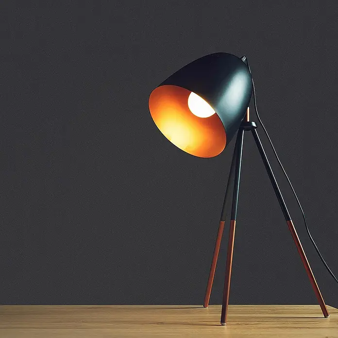 Tadessi Vliestapete M1-No.1906 - Schwarz, Uni, 10,05 x 0,53 m - Moderne schwarze Schreibtischlampe mit kupferfarbenen Stativbeinen, die warmes Licht auf einen Holztisch wirft.