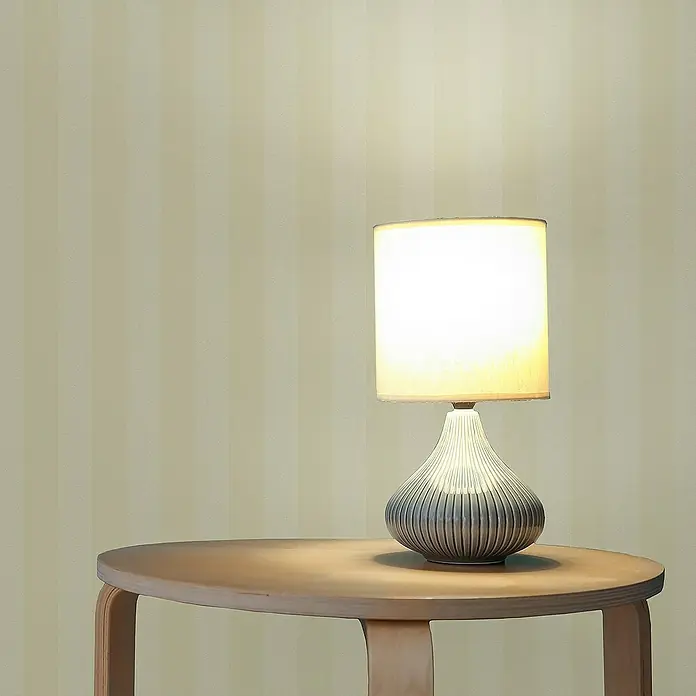 Tadessi Vliestapete M1-No.2944 - Gelb, Streifen, 10,05 x 0,53 m - Beleuchtete Tischlampe mit zylindrischem Schirm und geripptem, grauem Fuß auf einem hölzernen Beistelltisch.
