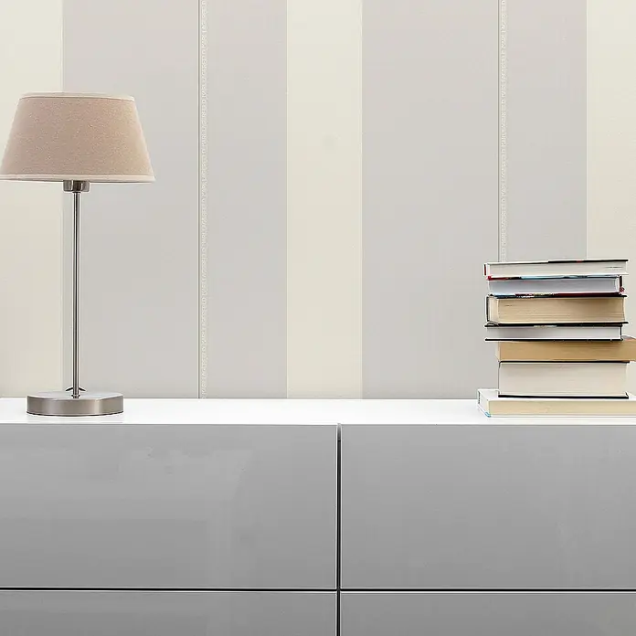 Tadessi Vliestapete M1-No.592 - Grau, Streifen, 10,05 x 0,53 m - Karl Lagerfeld Streifentapete in Hellgrau und Beige, dezent gebrandet, ergänzt ein modernes Interieur mit Lampe und Büchern.