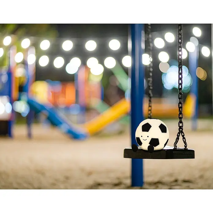 FHL easy LED-Nachtlicht Kalle - 0,4 W, L x B x H: 14 x 11,5 x 10,5 cm, Warmweiß, Fußball Kalle - Eine niedliche, sitzende Fußballlampe mit Gesicht und kleinen Beinen auf einer Schaukel im bunten Park, beleuchtet mit Lichterketten.