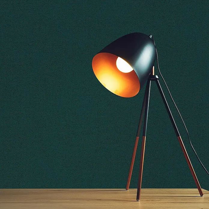 Tadessi Vliestapete M1-No.3174 - Grün, Uni, 10,05 x 0,53 m - Schwarze Tischlampe mit Kupfer-Innenseite und Stativbeinen auf Holzschreibtisch, strahlt warmes Licht ab.