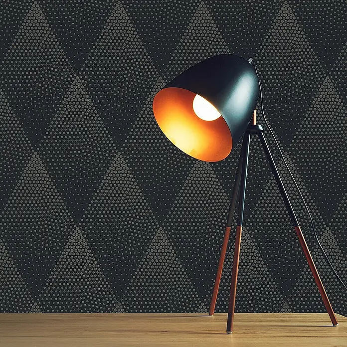 Tadessi Vliestapete M1-No.407 - Schwarz, Grafisch, 10,05 x 0,53 m - Eine stilvolle schwarze Tripod-Tischlampe mit warm leuchtender Glühbirne steht auf einer Holzoberfläche vor einer dunklen, gemusterten Wand.