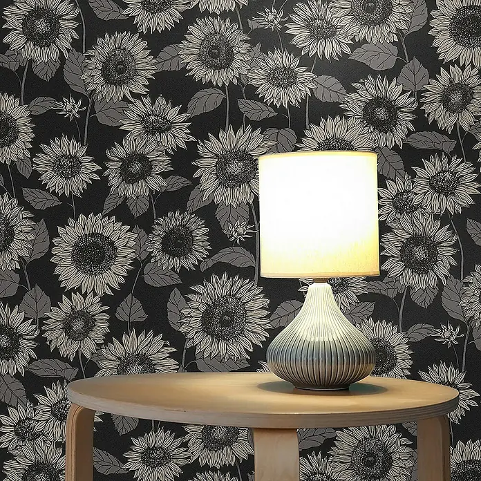 Tadessi Vliestapete M1-No.527 - Schwarz, Floral, 10,05 x 0,53 m - Eine stilvolle Tischlampe mit geripptem blau-grauem Keramikfuß und weißem Stoffschirm, auf einem runden Holztisch vor dunkler Sonnenblumen-Tapete.