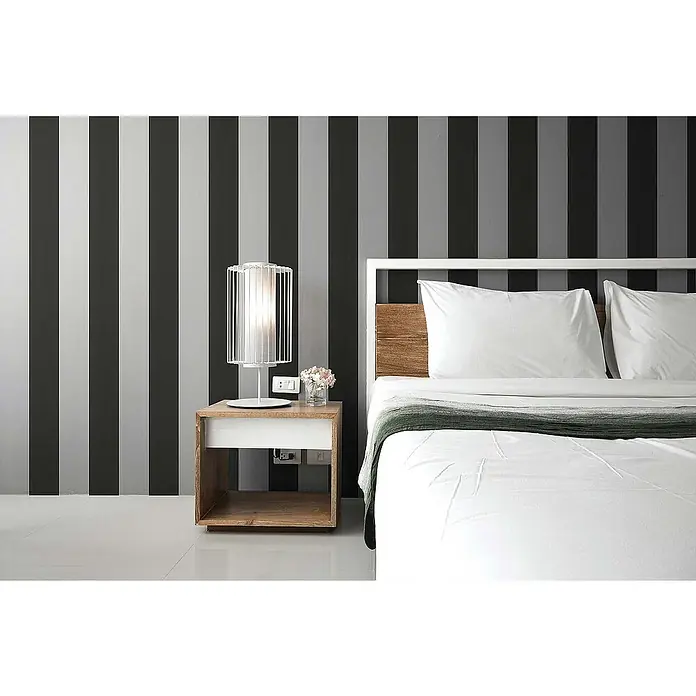 Tadessi Vliestapete M1-No.74 - Schwarz, Streifen, 10,05 x 0,53 m - Modernes Schlafzimmer mit Bett, Holz-Kopfteil, weißer Bettwäsche, Nachttisch aus Holz und weißer Lampe vor gestreifter Wand.