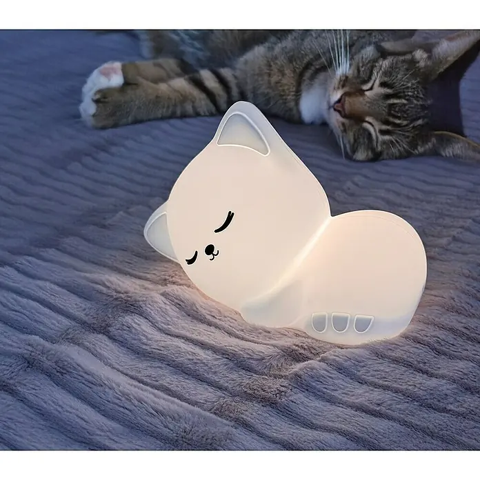 FHL easy LED-Nachtlicht Mia - 0,4 W, L x B x H: 16 x 8 x 11 cm, RGBW, Katze Mia - Eine niedliche, schlafende Katzenlampe leuchtet sanft auf einer weichen Decke, während eine echte Katze im Hintergrund schläft.