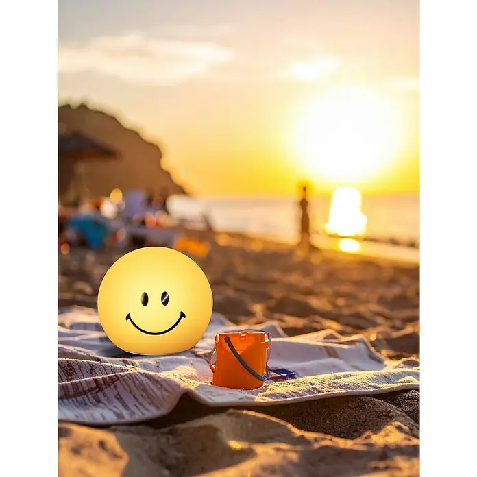 FHL easy LED-Nachtlicht GlowMoji - 0,3 W, L x B x H: 13 x 13 x 12 cm, Warmweiß, Smiley Yellow - Ein gelbes Smiley-Gesicht neben einem orangefarbenen Spielzeugeimer auf einem Strandtuch, mit einem wunderschönen Sonnenuntergang im Hintergrund.