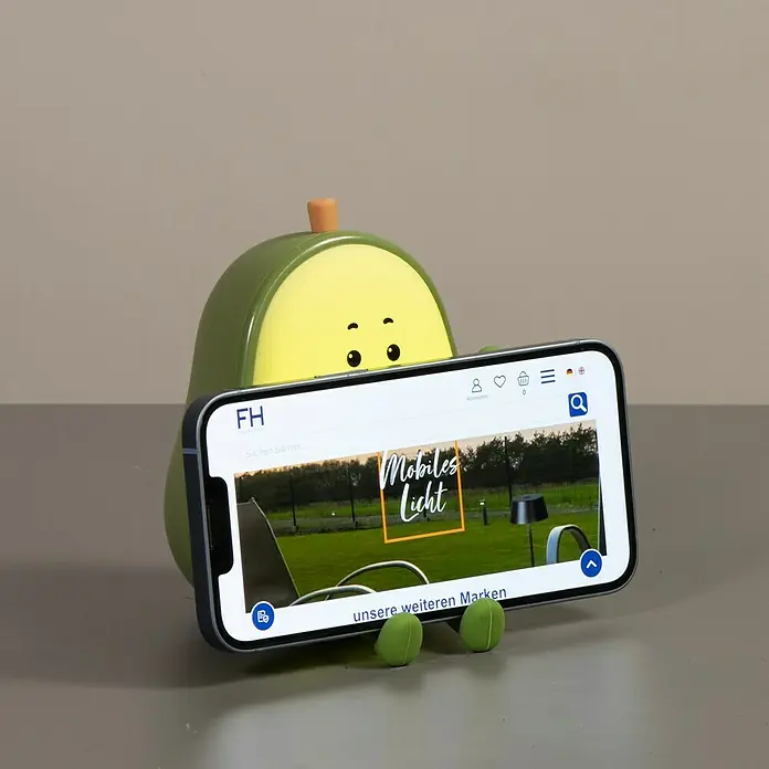FHL easy LED-Nachtlicht Bobi - 0,4 W, L x B x H: 13,5 x 9,5 x 13,5 cm, Warmweiß, Avocado Bobi - Das Bild zeigt einen Avocado-Handyhalter, der ein Smartphone hält. Auf dem Bildschirm des Handys ist eine Website zu sehen.