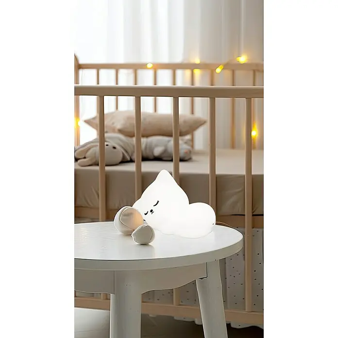 FHL easy LED-Nachtlicht Mia - 0,4 W, L x B x H: 16 x 8 x 11 cm, RGBW, Katze Mia - Ein schlafendes Wolken-Nachtlicht auf einem weißen Tisch neben einem Babybett mit Lichtern.