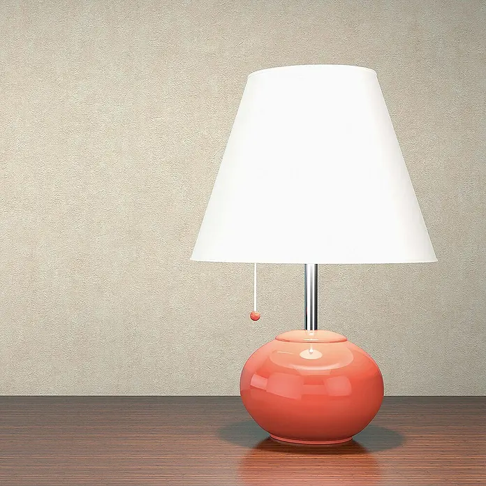 Tadessi Vliestapete M1-No.1815 - Creme, Steinoptik, 10,05 x 0,53 m - Glänzende korallfarbene Tischlampe mit weißem Schirm auf dunklem Holztisch vor beiger Wand.