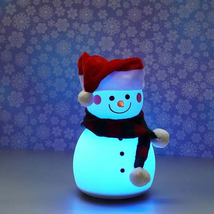 FHL easy LED-Nachtlicht Paule - 0,4 W, L x B x H: 11,6 x 11,6 x 22 cm, RGBW, Schneemann Paule - Das Bild zeigt einen leuchtenden Schneemann mit roter Mütze und Schal auf einem Tisch mit Schneeflocken im Hintergrund.