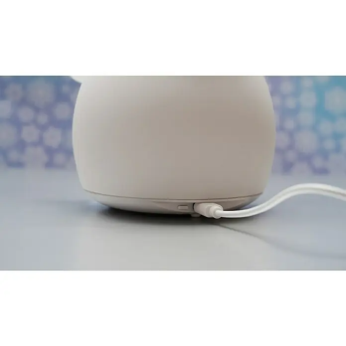 FHL easy LED-Nachtlicht Paule - 0,4 W, L x B x H: 11,6 x 11,6 x 22 cm, RGBW, Schneemann Paule - Nahaufnahme eines weißen Luftbefeuchters mit angeschlossenem Ladekabel. Das Gerät steht auf einer glatten Oberfläche vor einem verschwommenen Hintergrund.
