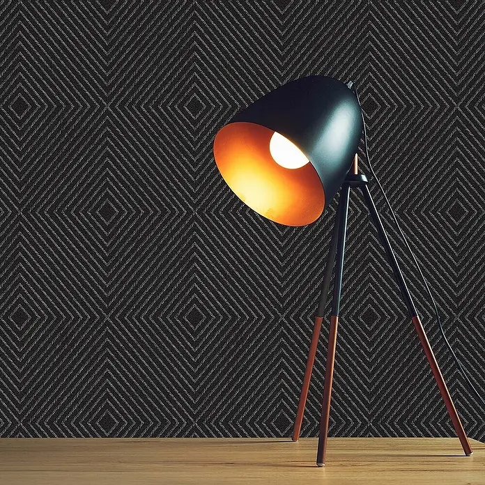 Tadessi Vliestapete M1-No.1587 - Schwarz, Grafisch, 10,05 x 0,53 m - Eine moderne schwarze Tischlampe mit glühender Glühbirne und Holzbeinen steht auf einer Holzoberfläche vor einer dunklen Wand mit dezentem geometrischem Muster.
