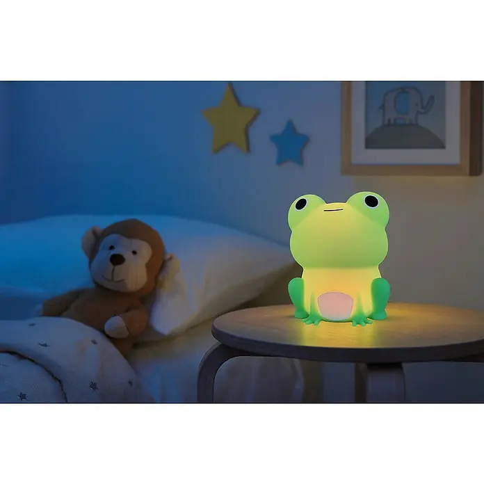 FHL easy LED-Nachtlicht Willi - 0,4 W, L x B x H: 9 x 8 x 11,5 cm, RGBW, Frosch Willi - Eine grüne Froschlampe steht auf einem Nachttisch neben einem Stofftier-Affen. Die Lampe strahlt ein sanftes, beruhigendes Licht aus.