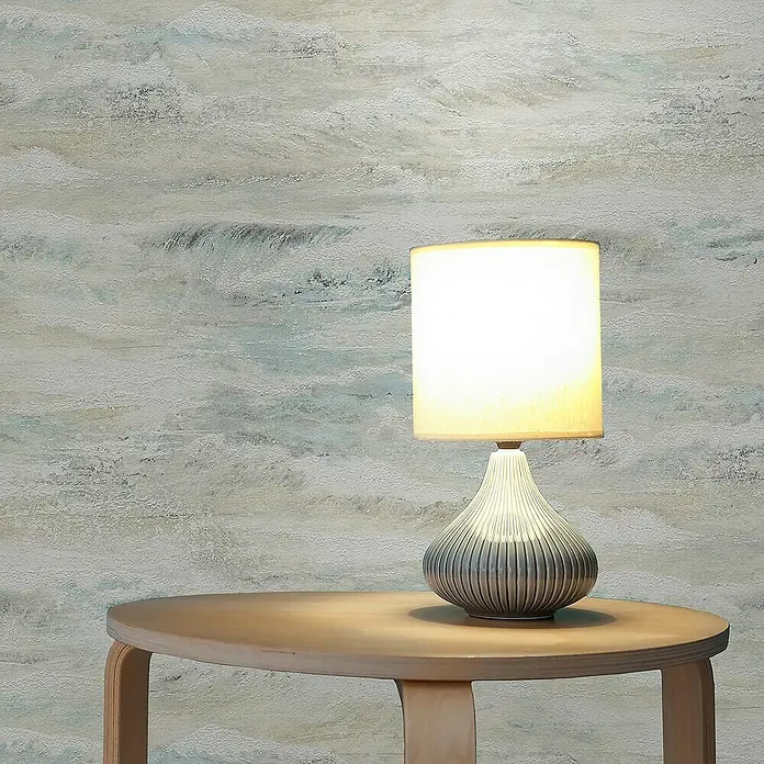 Tadessi Vliestapete M1-No.2573 - Grau, Streifen, 10,05 x 0,53 m - Eine Tischlampe mit hellem Stoffschirm und geriffelten grauem Sockel steht auf einem hellen Holztisch vor einer gemusterten Wand.
