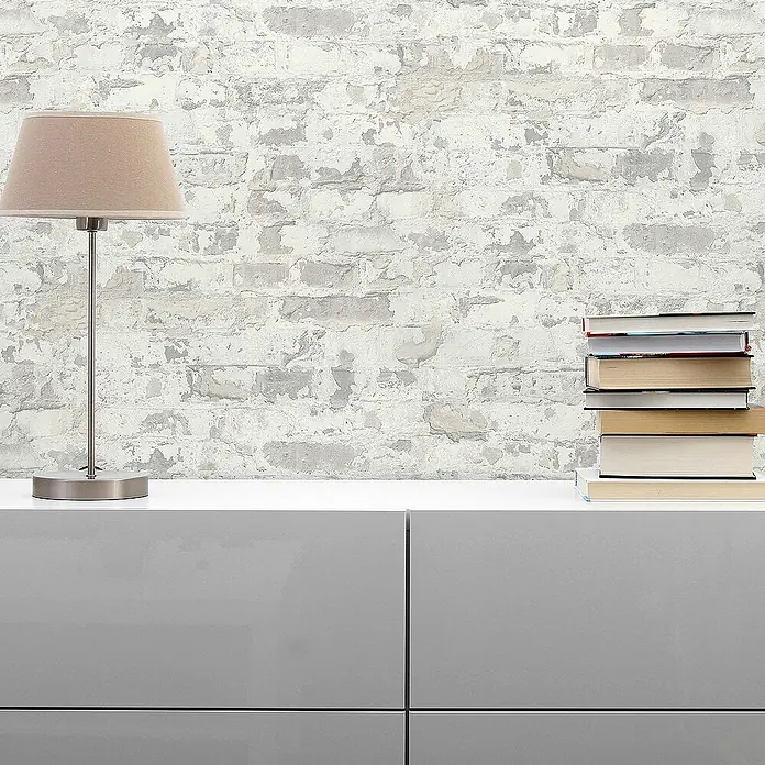 Tadessi Vliestapete M1-No.293 - Grau, Steinoptik, 10,05 x 0,53 m - Strukturierte weiße Ziegelstein-Tapete im Used-Look, dekoriert mit Tischlampe und Büchern auf einem Schrank.