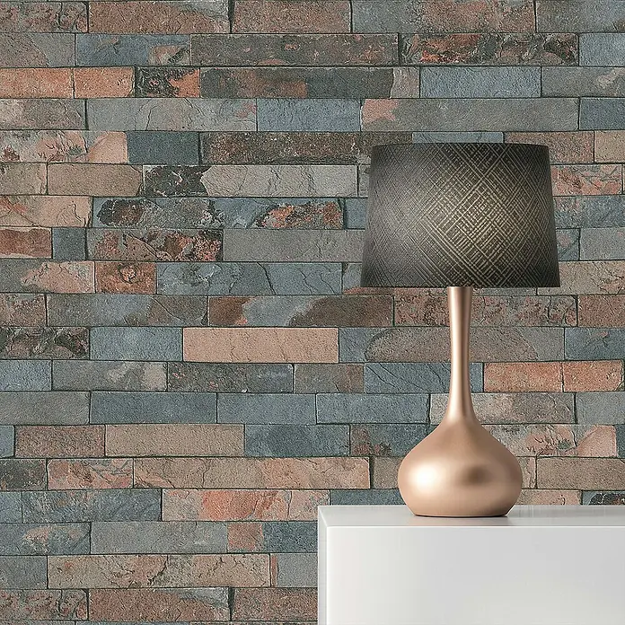 Tadessi Vliestapete M1-No.111 - Grau, Steinoptik, 10,05 x 0,53 m - Tischleuchte mit dunklem, gemustertem Schirm und metallic-roségoldenem Fuß vor einer bunten Ziegelwand.