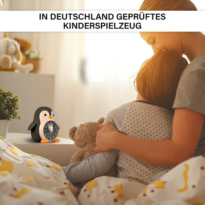 MegaLight Nachtlicht Wecker Poppy Penguin - Schwarz, Weiß, 14,8 x 13,8 cm - Die Mutter umarmt ihr Kind im Bett, im Vordergrund ein pinguinförmiger Kinderwecker. Geprüftes Kinderspielzeug aus Deutschland.