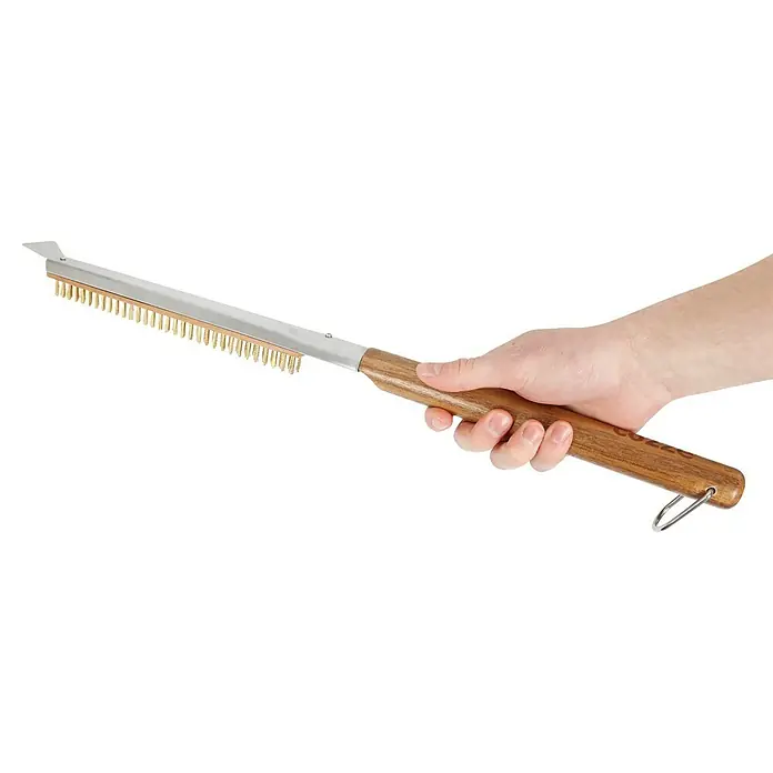 Cozze Grillbürste - Länge: 63 cm - Die Hand hält eine Grillbürste mit Holzgriff und goldenen Borsten zum Reinigen von Grillrosten. Praktisches Grillzubehör für saubere Grills.