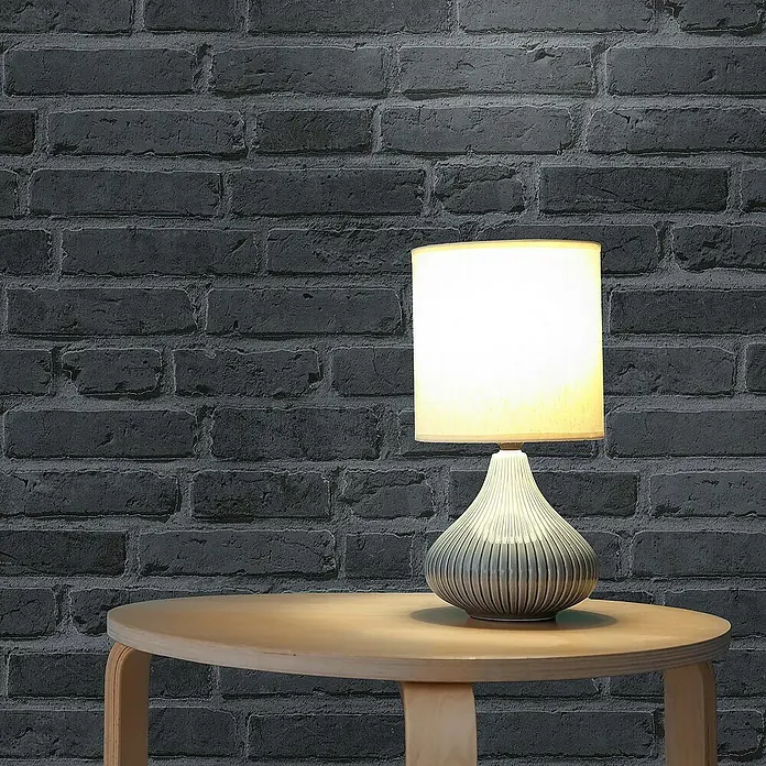Tadessi Vliestapete M1-No.890 - Schwarz, Steinoptik, 10,05 x 0,53 m - Eine Tischlampe mit beigem Schirm und grau-blauem Keramikfuß steht auf einem hellen Holztisch vor einer dunklen Ziegelwand.