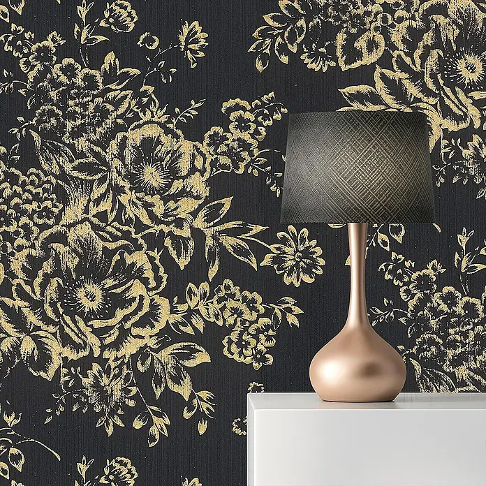 Tadessi Vliestapete M1-No.2169 - Schwarz, Floral, 10,05 x 0,53 m - Roségoldene Tischleuchte mit dunkelgrauem, gemustertem Lampenschirm, auf weißer Oberfläche vor schwarz-goldener Blumentapete.