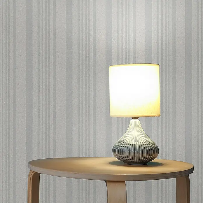 Tadessi Vliestapete M1-No.3611 - Weiß, Streifen, 10,05 x 0,53 m - Eine stilvolle Tischlampe mit geripptem, grau-blauem Keramiksockel und cremefarbenem zylindrischem Schirm, die warmes Licht auf einen runden Holztisch wirft.