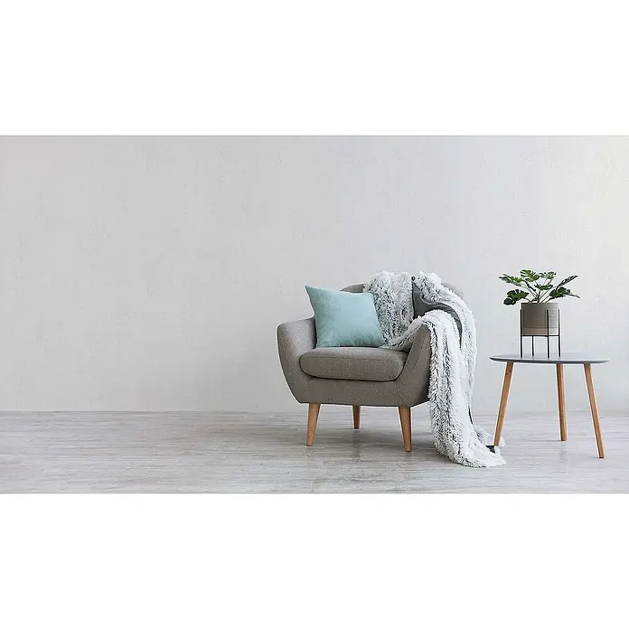 Tadessi Vliestapete M1-No.587 - Weiß, Steinoptik, 10,05 x 0,53 m - Ein grauer Sessel mit blauem Kissen und flauschiger Decke, daneben ein Beistelltisch mit Pflanze.
