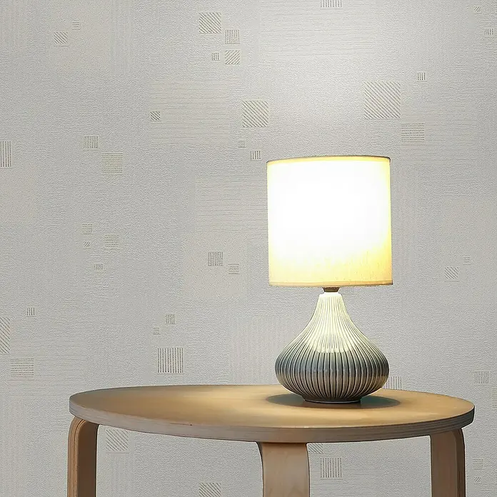 Tadessi Vliestapete M1-No.2444 - Weiß, Sonstiges, 10,05 x 0,53 m - Eine Tischlampe mit weißem Schirm und grauem, geriffeltem Fuß steht auf einem hellen runden Holztisch und spendet warmes Licht.