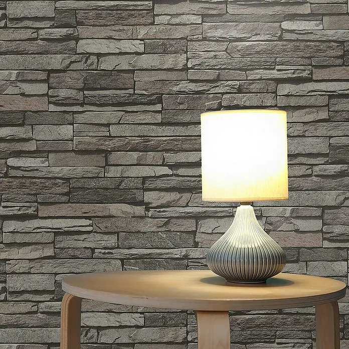 Tadessi Vliestapete M1-No.8 - Schwarz, Steinoptik, 10,05 x 0,53 m - Beleuchtete Tischlampe mit hellem Schirm und geripptem, grauem, birnenförmigem Sockel auf Holztisch vor Ziegelwand.