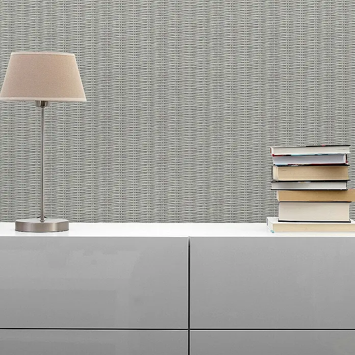 Tadessi Vliestapete M1-No.1653 - Grau, Holzoptik, 10,05 x 0,53 m - Graue Wandverkleidung mit gewebter, rattanähnlicher Textur, in einem Raum hinter einer Lampe und Büchern angebracht.