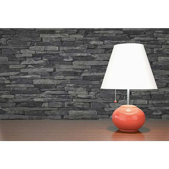 Tadessi Vliestapete M1-No.2 - Schwarz, Steinoptik, 10,05 x 0,53 m - Orange Tischlampe mit weißem Schirm auf Holzfläche vor dunkler Steinwand.