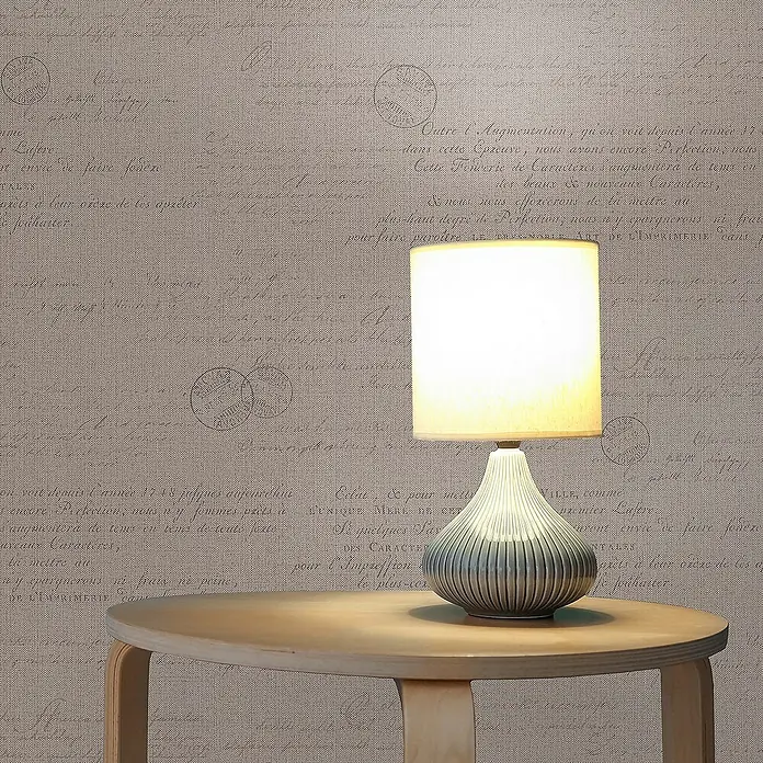 Tadessi Vliestapete M1-No.1513 - Beige, Sonstiges, 10,05 x 0,53 m - Eine Tischlampe mit cremefarbenem Schirm und geriffelter Keramikbasis steht auf einem runden Holztisch vor einer gemusterten Tapete mit Vintage-Schrift.