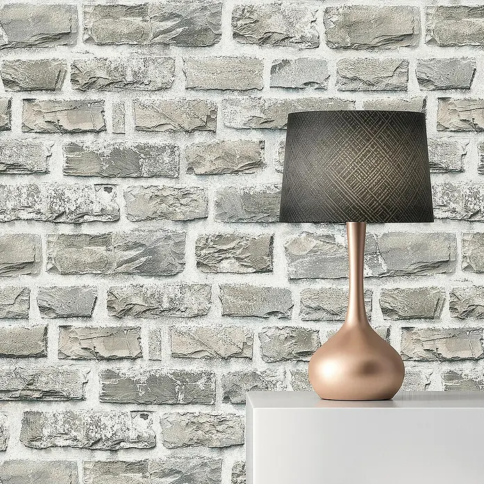 Tadessi Vliestapete M1-No.2635 - Grau, Steinoptik, 10,05 x 0,53 m - Eine moderne Tischlampe mit Kupfersockel und dunklem Musterschirm auf einer weißen Fläche vor einer Steinziegelwand.