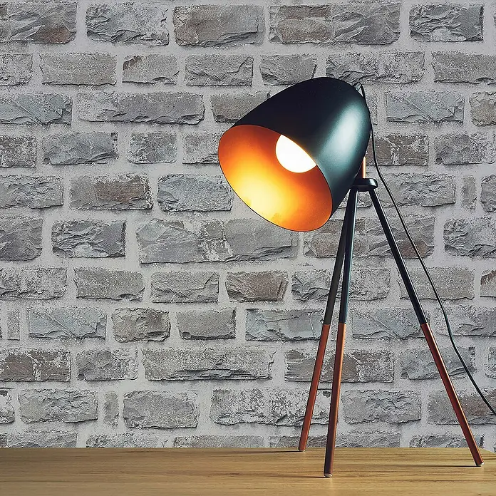 Tadessi Vliestapete M1-No.161 - Mehrfarbig, Steinoptik, 10,05 x 0,53 m - Eine stilvolle schwarze Metall-Dreibein-Stehlampe mit orangefarbener Innenseite, auf einem Holztisch, die eine Steinmauer beleuchtet.